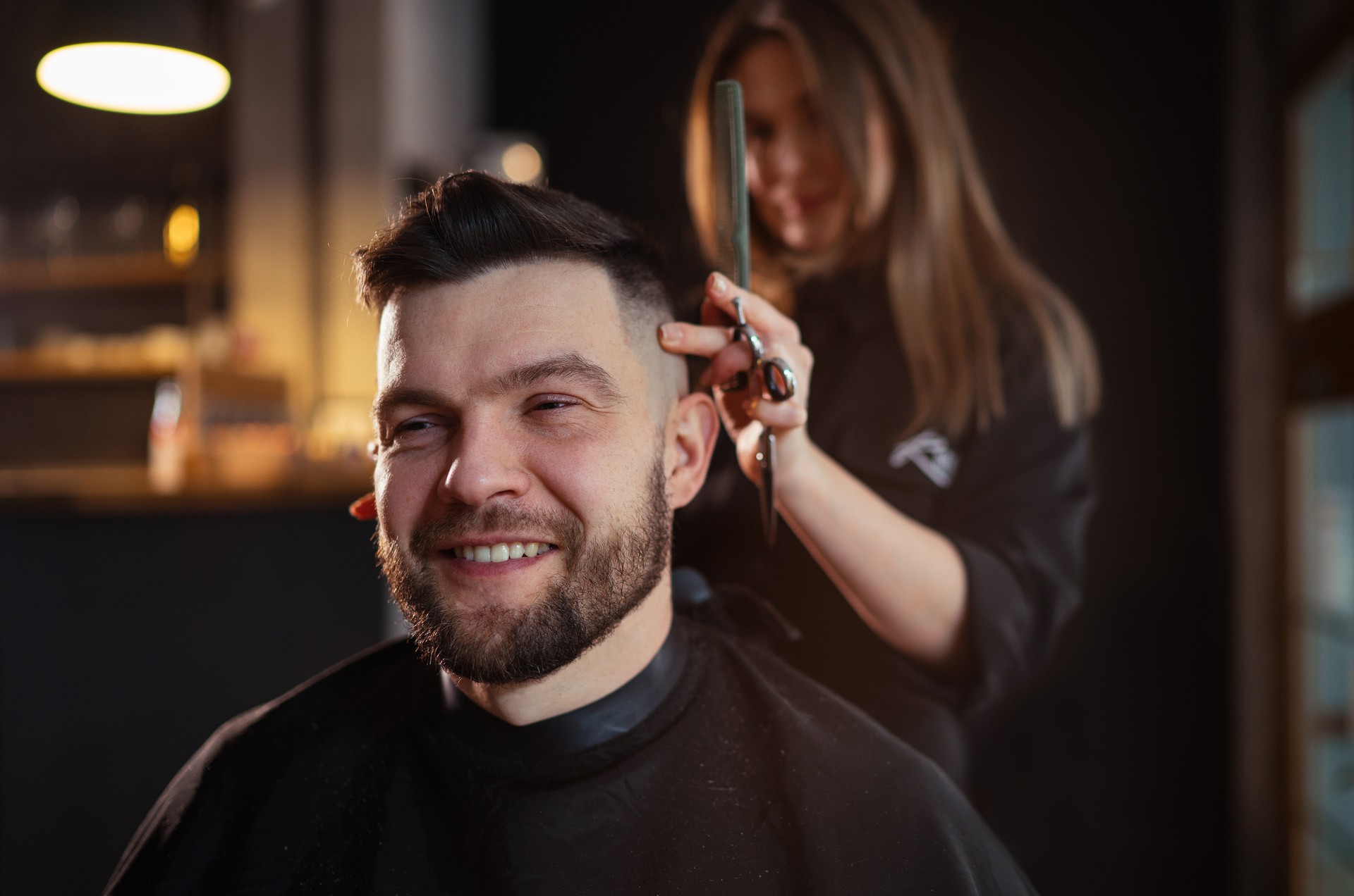 Portrait d’homme barbu souriant tout en coupe de coiffure dans un salon de coiffure par une jeune coiffeuse. Intérieur moderne de salon de coiffure de style noir à faible luminosité. Concept de petite entreprise de service de soins capillaires